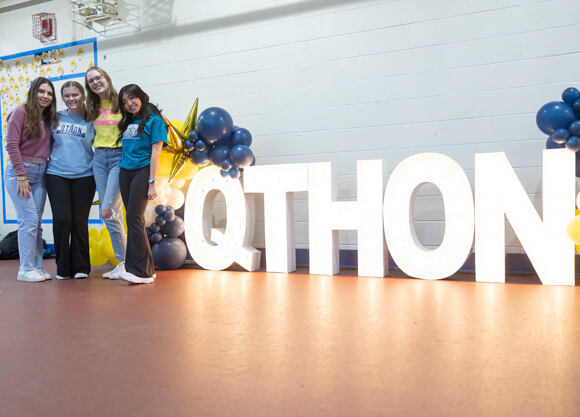 Four students pose for a photo in front of a giant light up sign that reads QTHON